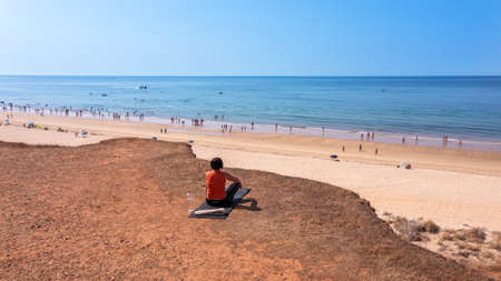 A middle-aged woman, during a vacation in Portugal, sits on a rug on a cliff, in front of the sea and meditates. High quality photoの写真素材