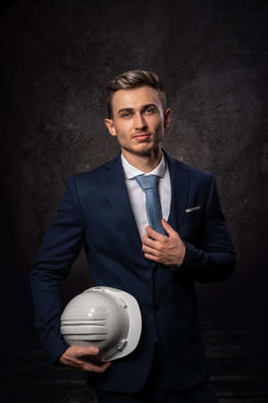 Young caucasian businessman architect engineer, with a white protective helmet in hand for construction. Close-up, with a smart faceの写真素材