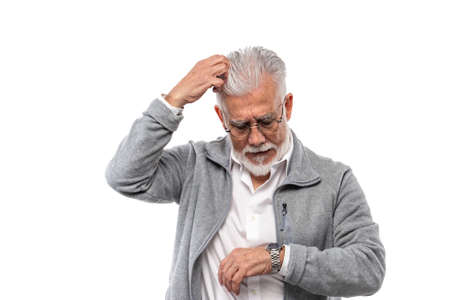 An elderly stylish man, gray-haired and with a beard on a white background, scratches his head and looks at his watch. The concept of punctuality and disciplineの写真素材