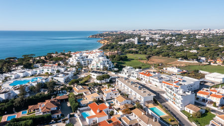 Aerial view of Olhos de Agua with whitewashed houses, resort pools and coastline leading to Falesia cliffs under clear blue sky in Albufeira, Algarve, Portugal. Concept of sunny beachside tourismの写真素材