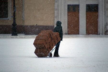 man carrying a lot of cardboard boxes on a trolley on a snow-marked road during a snowstormの写真素材