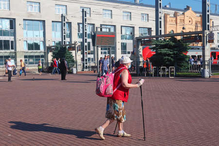 June 18, 2020, Russia, St. Petersburg, an elderly woman in a medical mask with a backpack and sticks for walking and a white in hand is walking along the platform to the trainのeditorial素材