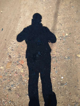 shadow of a man with a cap on his head on a surface of sand and stones on a sunny summer dayの写真素材