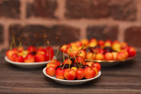 three servings of different varieties of sweet cherries on plates on a wooden table against a brick wall backgroundの写真素材
