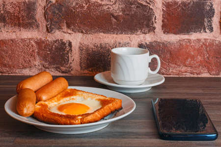 coffee, sausages and a hot egg sandwich for breakfast and a mobile phone, with a brick wall reflected on the screen on a wooden tableの写真素材