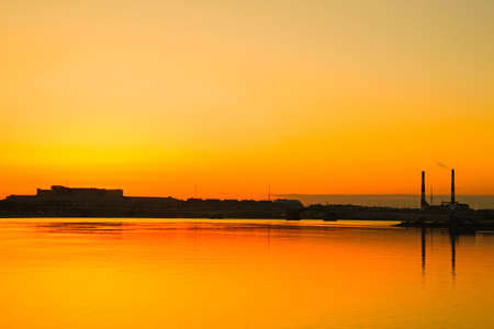 silhouettes of industrial buildings and factory pipes on the shore, reflected in the water at sunset