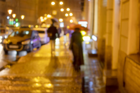blurred silhouettes of people with umbrellas walking through the evening city in the rainの写真素材