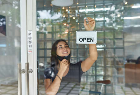 Beautiful European girl removing a mask, hangs a sign on the store door (open) after a cowid-19...