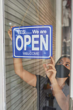 Beautiful European girl in a mask hangs a sign on a glass door of a clothing store (open) after a...