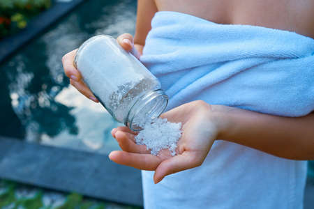 A girl in a white towel pours sea salt into her hand from a glass jar against the backdrop of a pool for body scrub.の写真素材