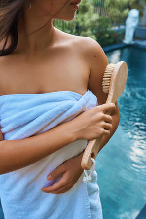 A girl in a white towel massages the skin of her hands with a brush against the background of the pool. Dry massage brush. Zero waste conceptの写真素材