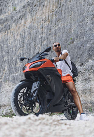 A young guy with glasses sits on a red sports motorcycle against the background of rocks