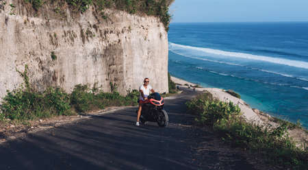 A young guy with glasses sits on a red sports motorcycle against the backdrop of the seaの写真素材