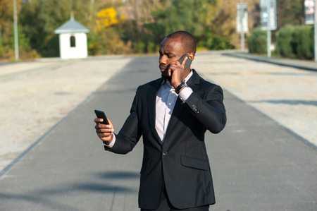 a black man in a business suit standing outdoors and uses two phonesの写真素材
