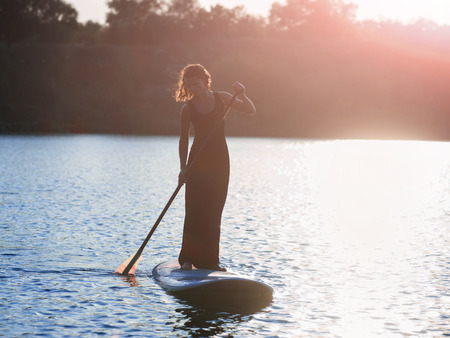 Beautiful woman in a black dress on stand up paddle board,sunset.の写真素材