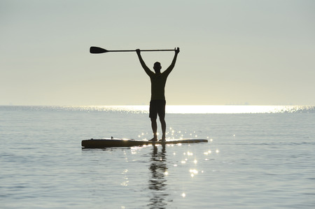 SUP silhouette of athletic man standing with a paddle on the surfboard at sunset stand up paddle boardingの写真素材