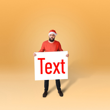 Full length portrait of surprised man in santa hat demonstraiting big card with inscription, isolated on orange backgroundの写真素材