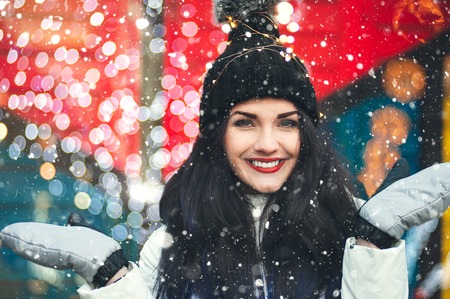 Winter holidays concept. Close up portrait of beautiful woman in warm hat and mittens. She is smiling and standing under snowfallの写真素材