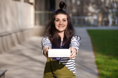 Smiling woman with funny hair bun holding white present box in outstretched hands with city street on backgroundの写真素材