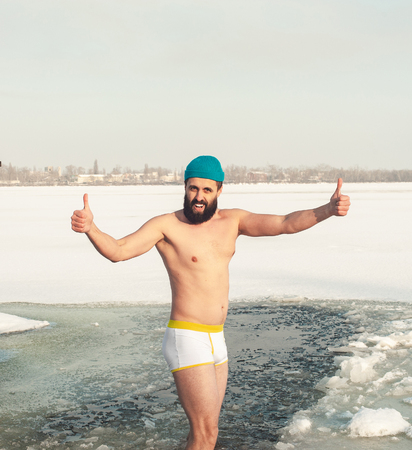 bearded man in hat swimming in the winter lake in the ice hole, ice hole swimming, selfie before diving into the ice hole, thumb upの写真素材
