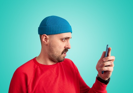 Perplexity facial expression concept. Side view of stylish mustache man in bright clothes holding a phone in front of him and looking at it with disbeliefの写真素材