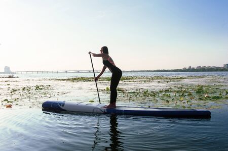 Young athletic woman doing fitness on a board with an oar on a lake.