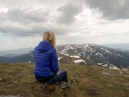 A blonde girl in a jacket sits with her back on a high mountain. Against the backdrop of clouds and high mountainsの写真素材