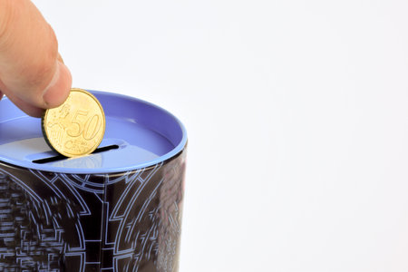 A hand holds 50 euro cents over a piggy bank on a white background. On the right is an empty space...