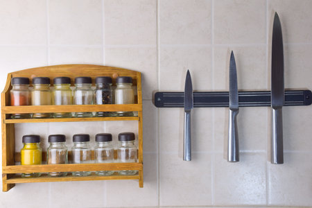 A set of jars with different spices and a set of three steel knives hang on the ceramic wallの写真素材