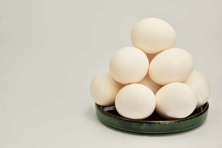 A pile of white chicken eggs lies in a green ceramic saucer. There is a place on the left for an...