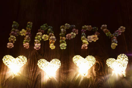 On a wooden background, the word HAPPY is spelled out in large letters of multicolored flowers. Glowing hearts are laid out in a line from belowの写真素材
