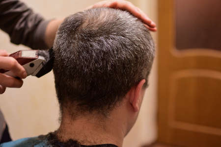 A grown man is trimmed with a clipper and sits with his back to the camera. Perhaps at home in a...