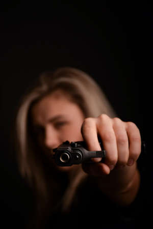 A young girl takes aim at the frame from a pistol, which she holds with both hands. Only a gun is in focus, and a girl on a blurry dark backgroundの写真素材
