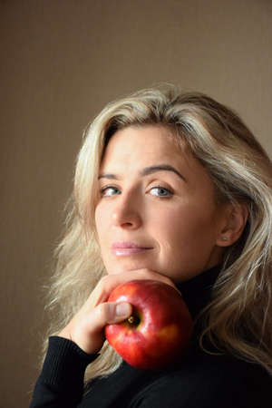 A beautiful blonde girl with long hair looks at the camera and holds a red apple with her hand....