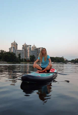 A beautiful woman floats on a SUP paddle sitting, smiling and relaxing. Multi-storey buildings in...