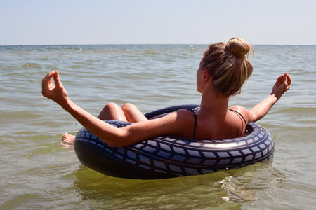 A young beautiful woman on a lifebuoy swims in the sea and holds her hands in a zen pose. back view