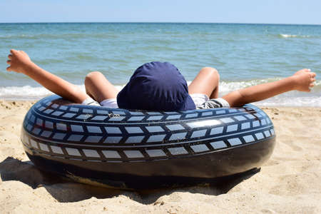 A boy in a cap lies on the sea sandy beach in a rescue inflatable ring with his arms outstretched. back viewの写真素材