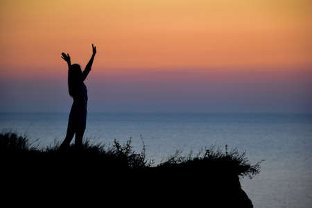 Silhouette of an unrecognizable woman with outstretched arms on a hill against the background of the...