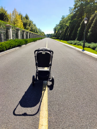 A baby carriage stands on a long asphalt road in perspective between a decorated fence and a park...