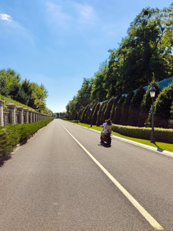 A boy and a girl are riding an electric scooter in perspective on a long asphalt road in a city park during the dayの写真素材