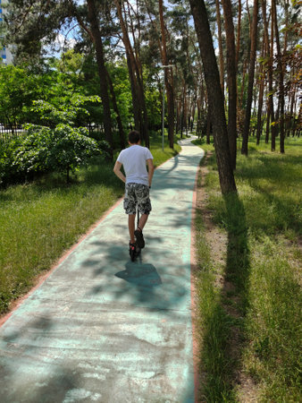 A teenage boy rides a scooter along a winding path in the park between trees. rear view