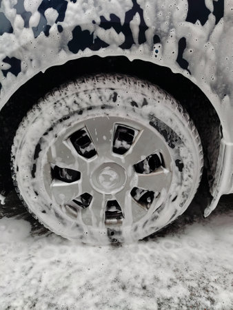 Car wheel in white washing foam with bubbles at car wash close-up. Foam flows down the sides of the car onto the asphaltの写真素材