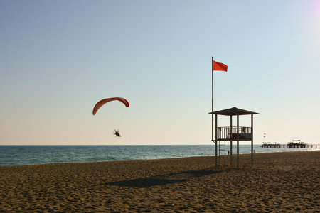 A paraglider flies over the evening sea beach. There is a sea rescue tower on the beach. The sky is clear and blueの写真素材