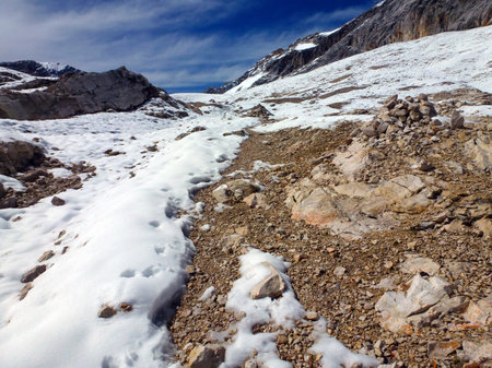 Scattered stone cobblestones on a snowy mountain peak against a background of a blue sky with light clouds and other mountains in the distanceの写真素材