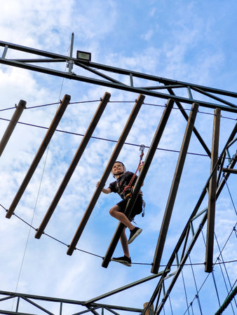 A teenager in sports equipment is crossing a suspended suspension rope bridge against the background of a blue sky. View from belowの写真素材