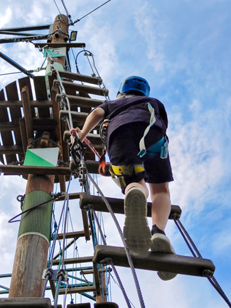 A teenage Caucasian boy in a rope amusement park climbs on a simulator against a blue sky. View from belowの写真素材