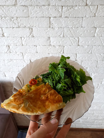 Homemade pie and spinach leaves on a plate against a brick wall in the kitchen. A female hand holds a plateの写真素材