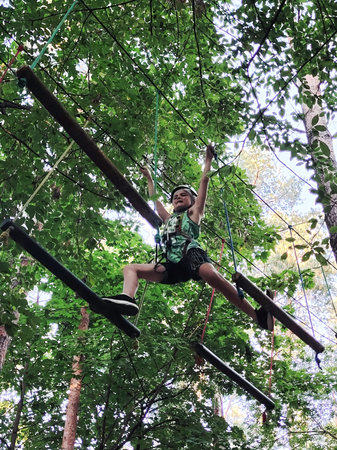 A teenage boy in a safety harness crosses a ropeway in an amusement park. View from below against a...