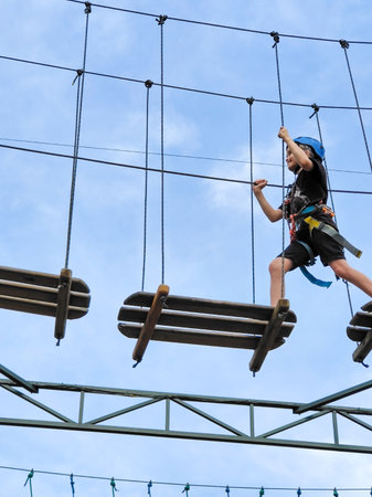 A teenage boy in a safety harness crosses a suspension bridge on a wooden obstacle course in an amusement park. Summer sportsの写真素材
