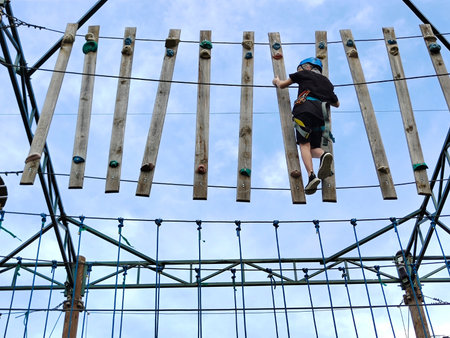 A teenage boy crosses between towers on a rope bridge in an amusement park. View from below against...
